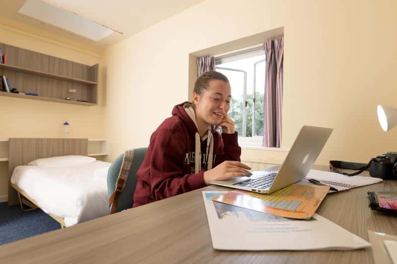 Teenage male student sitting at his desk in his university college bedroom, smiling and looking at his laptop
