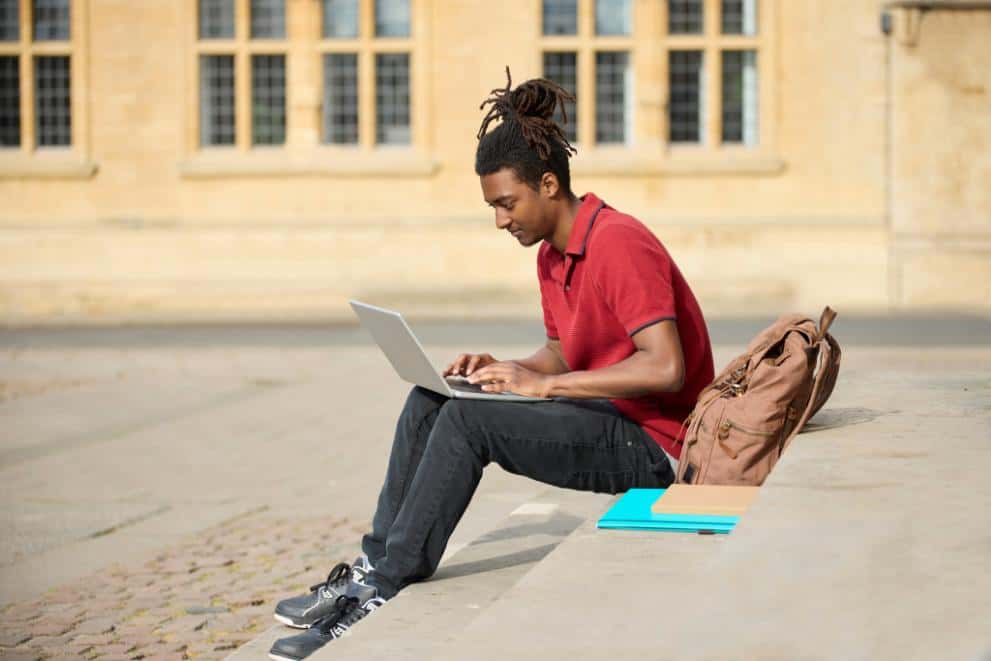 An adult summer school student sitting outside a university college building with his laptop